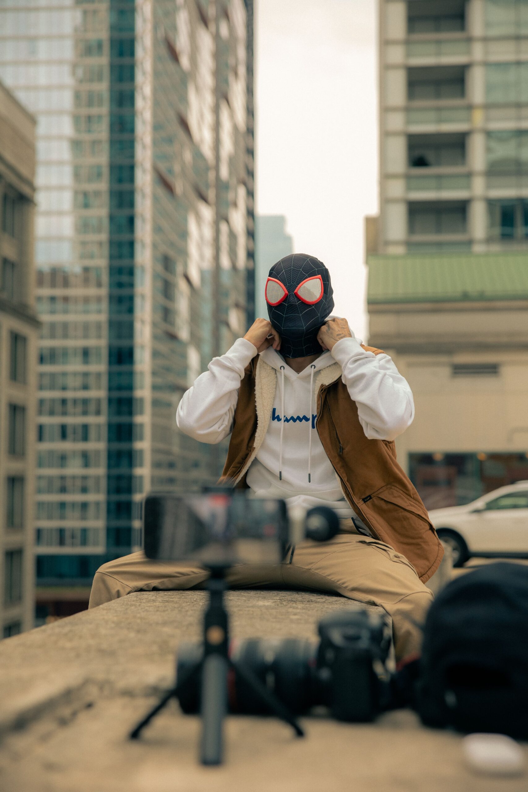 Person in superhero mask sitting on urban rooftop with camera equipment.