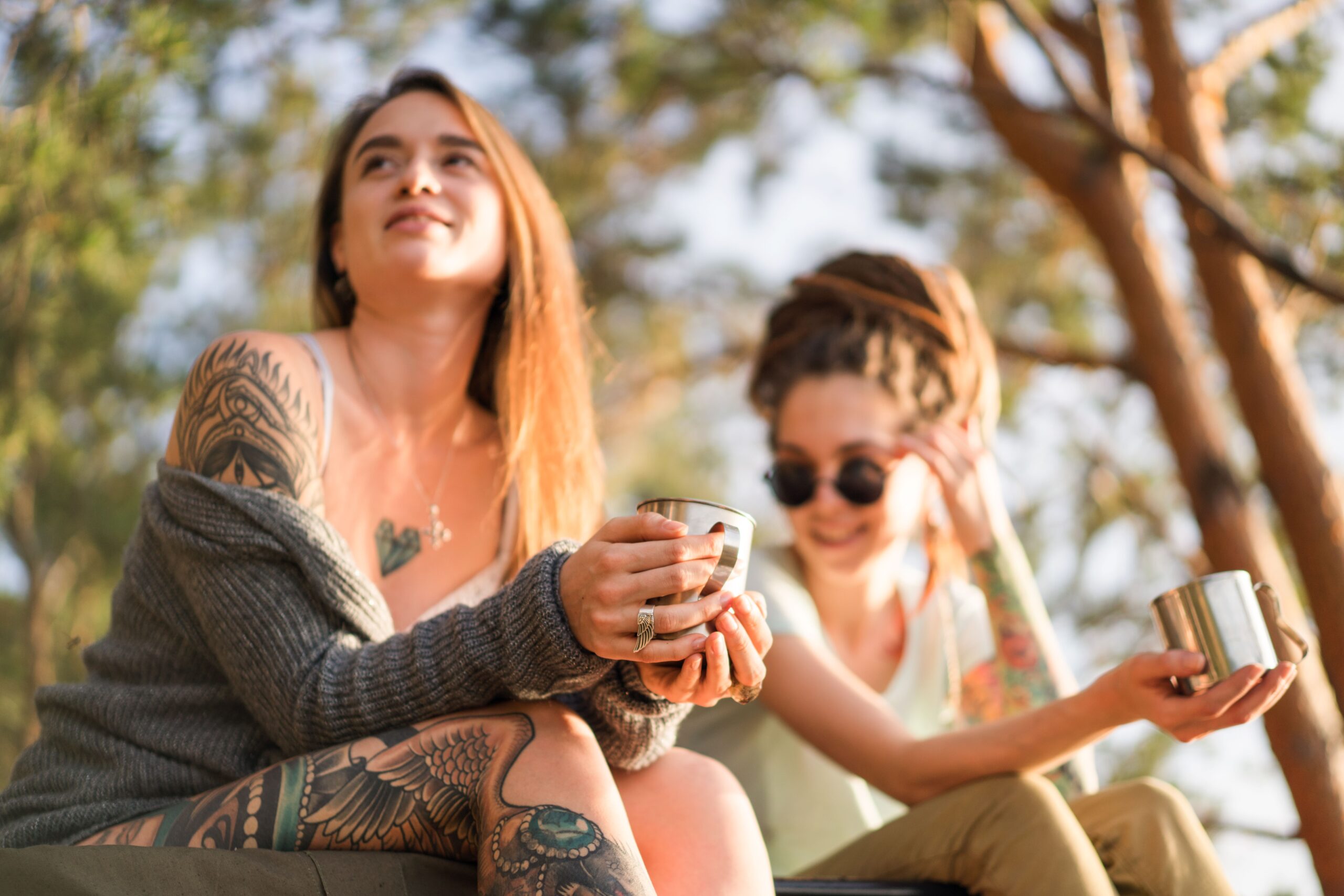 Two women with tattoos enjoying coffee outdoors, showcasing friendship and relaxation.
